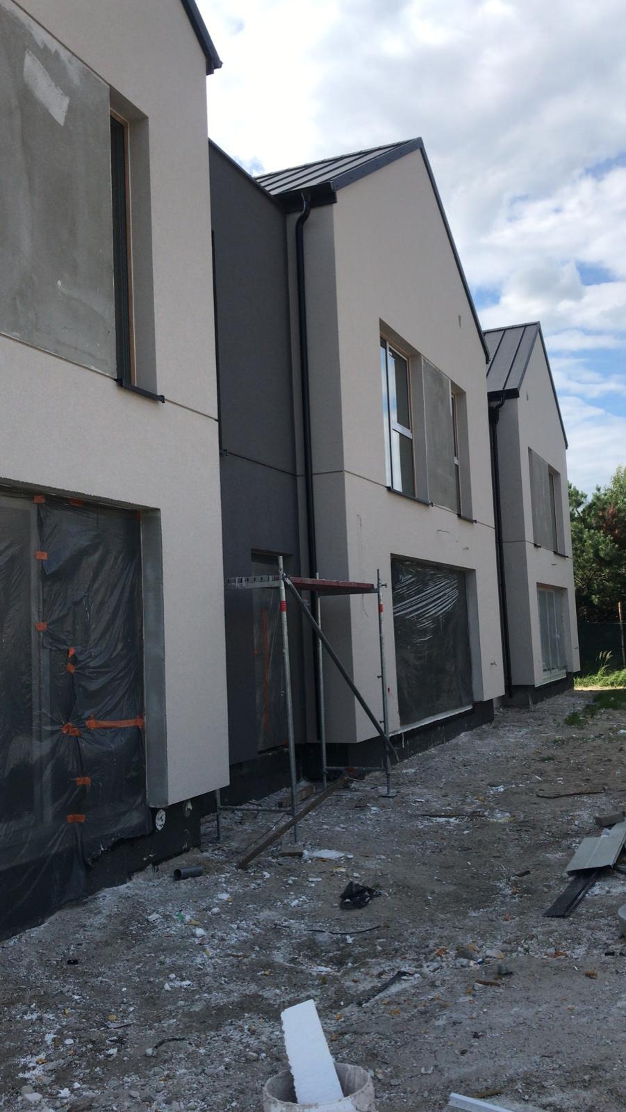 Trzy segmenty bliźniaka w trakcie wykończenia elewacji, z widocznym rusztowaniem i zabezpieczeniami okien folią, teren wokół w stanie surowym.