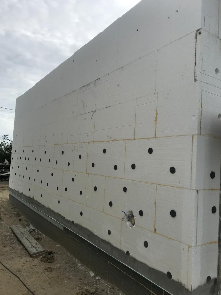 Ściana budynku w trakcie ocieplania styropianem, z widocznymi czarnymi kołkami mocującymi i otworem na instalację, na tle pochmurnego nieba.