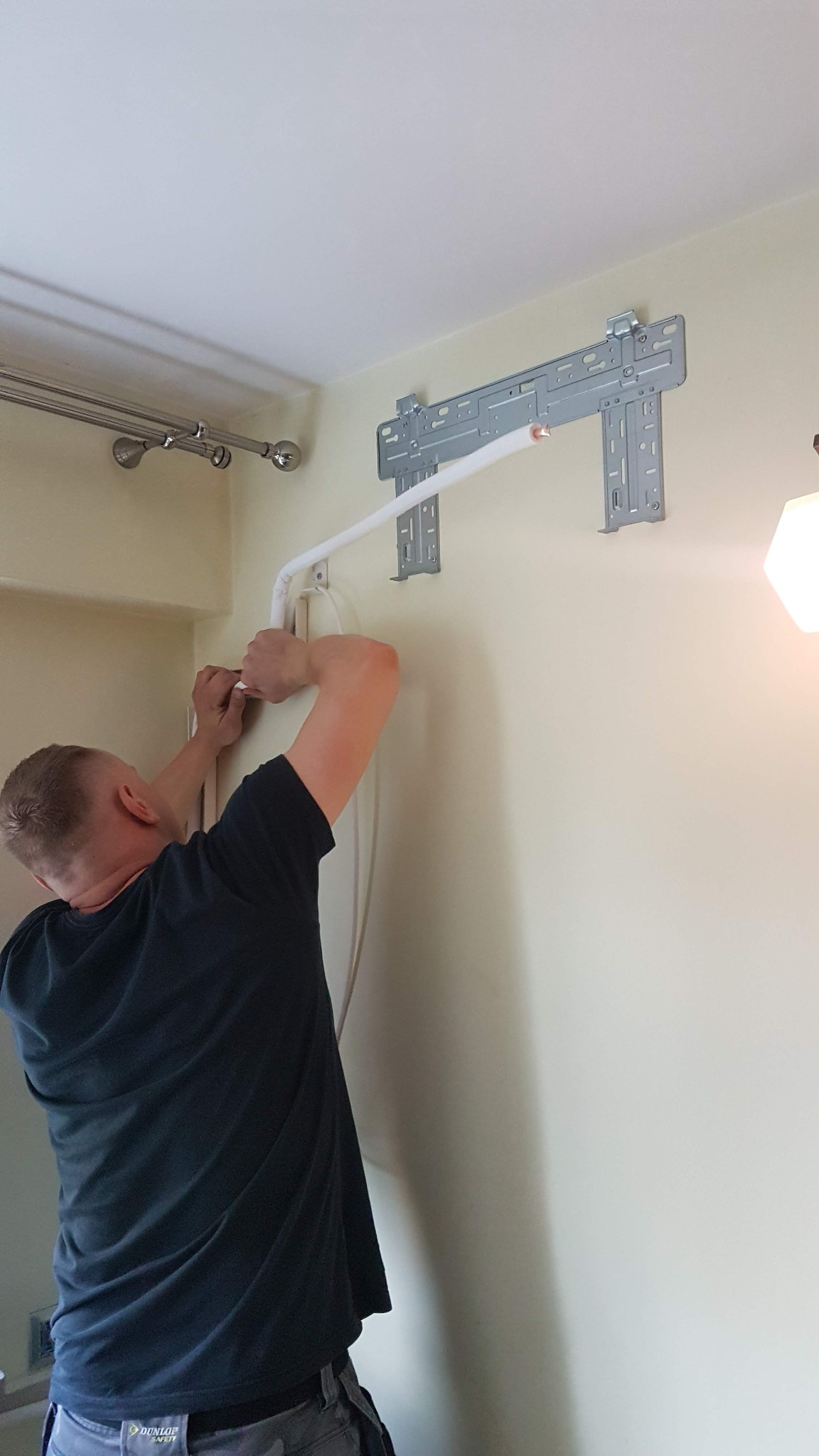 Instalacja klimatyzacji: monter podłącza rurę do uchwytu na ścianie w jasnym pomieszczeniu, widoczny fragment instalacji elektrycznej.