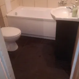 Biała wanna, umywalka z szafką i toaleta na ciemnobrązowych płytkach w wykafelkowanej łazience, widok zza framugi drzwi.