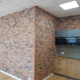 Wykończone wnętrze biura z elementami kuchni: ściana z cegły dekoracyjnej, drewniane szafki kuchenne oraz szare szafki wiszące z zabudowanym piekarnikiem i mikrofalówką, widoczna wentylacja sufitowa.