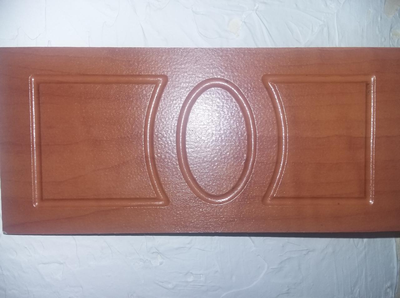 Lakierowany panel meblowy z wytłoczonym wzorem geometrycznym (owal i prostokąty) na tle białej, teksturowanej ściany. Ciepła tonacja brązu i beżu.