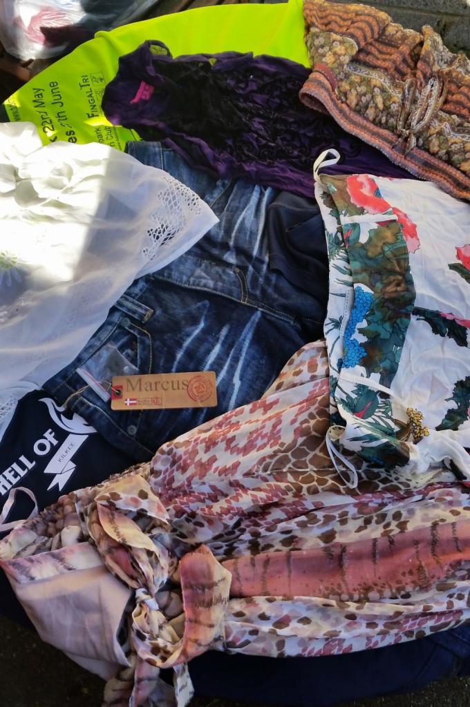 Stos różnorodnych ubrań, w tym dżinsy z metką 'Marcus' z flagą Danii, bluzka z motywem kwiatowym, koszulka z napisem 'HELL OF KILKEE', oraz inne tekstylia o różnych wzorach i fakturach.