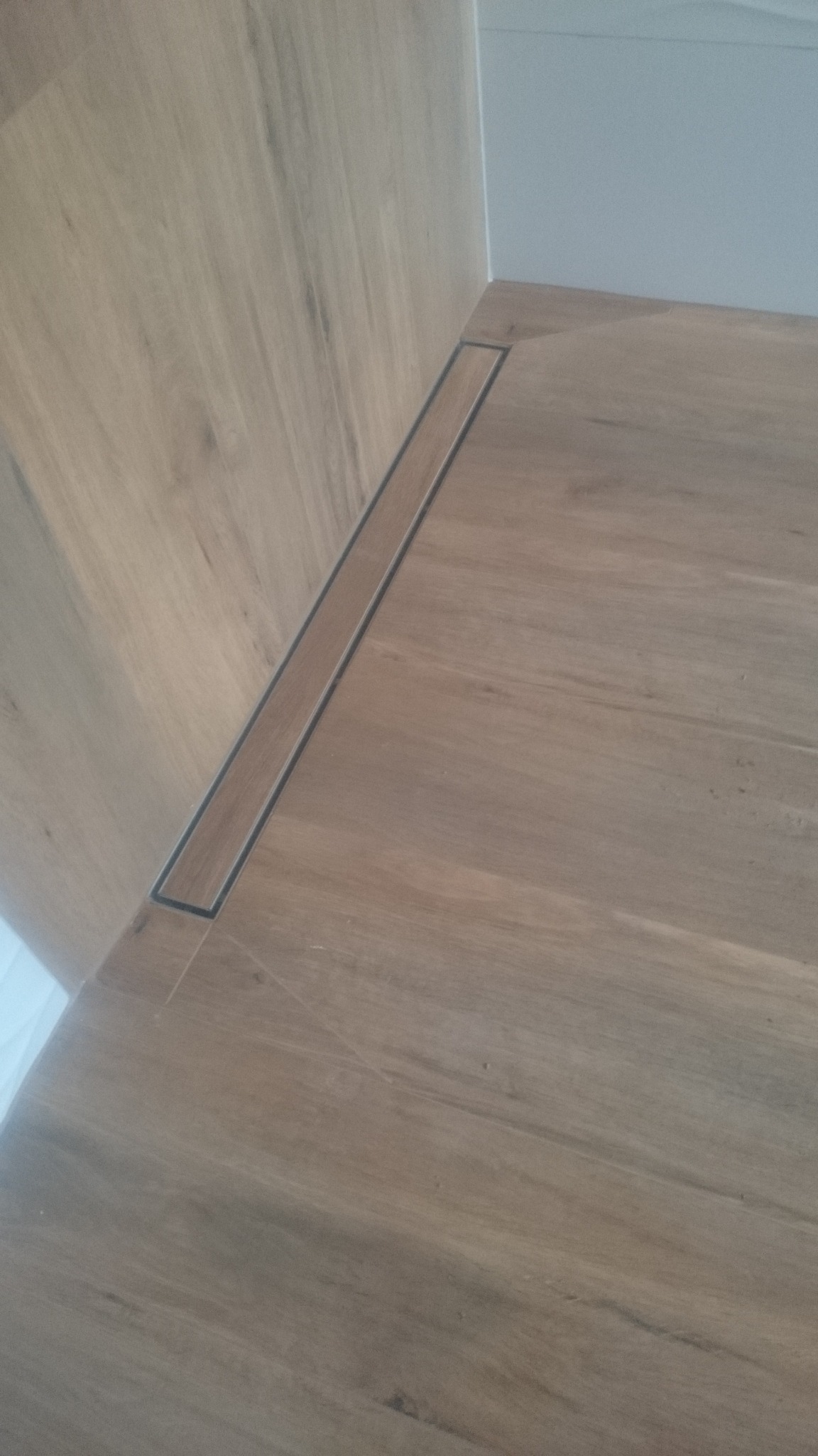 Ukończona podłoga prysznicowa z odpływem liniowym, wyłożona płytkami imitującymi drewno, z widocznym fragmentem ściany.