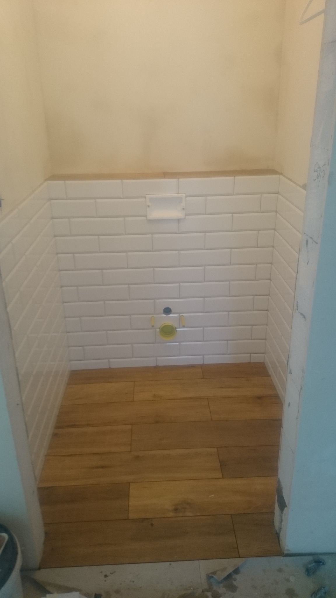 Niewielka łazienka w trakcie remontu: ściany wyłożone białymi, prostokątnymi płytkami, na podłodze ułożone płytki imitujące drewno, widoczne przygotowane przyłącze kanalizacyjne pod muszlę klozetową.