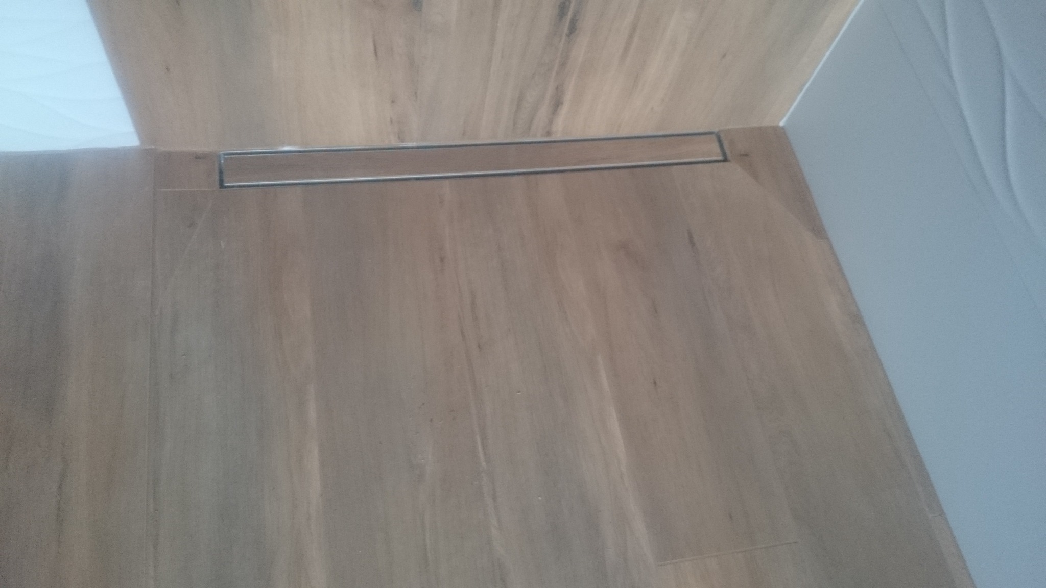 Fragment łazienki z drewnopodobnymi płytkami podłogowymi i odpływem liniowym w podłodze, widoczna również fragment białej ściany z dekoracyjną fakturą.