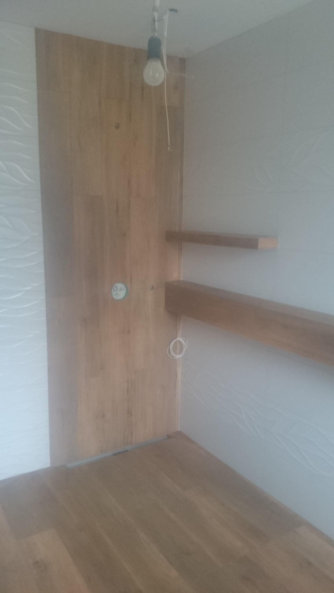 Wnętrze łazienki w trakcie wykańczania: drewnopodobne płytki ścienne i podłogowe, białe płytki z falistym wzorem na ścianie, drewniane półki, wisząca żarówka.