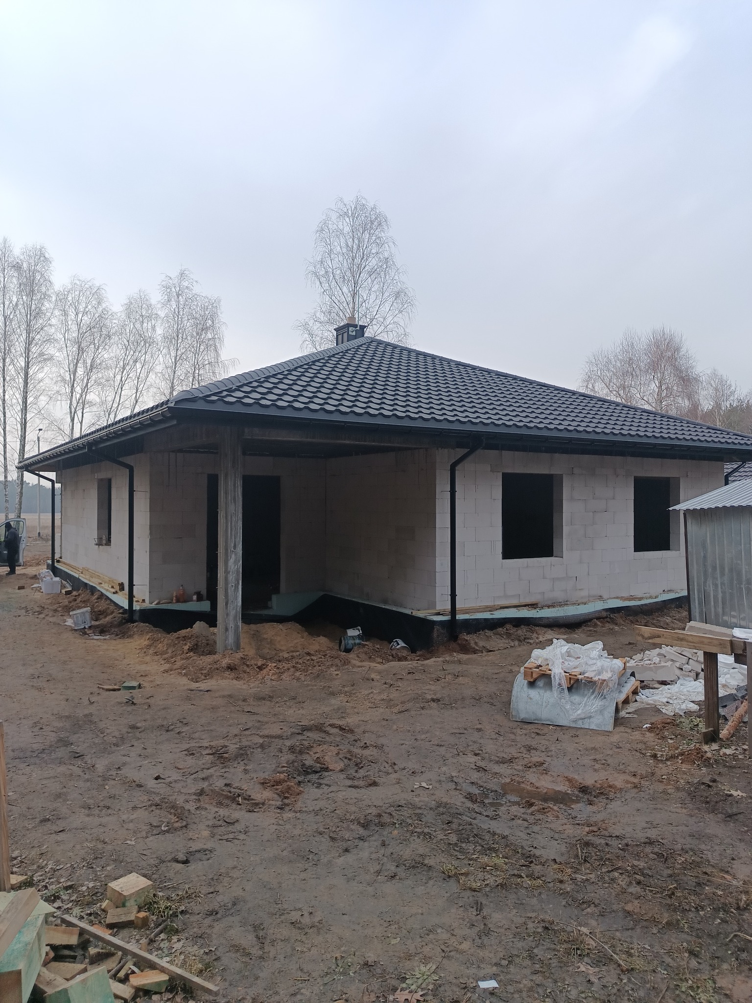 Niedokończony dom z białego pustaka z czarnym dachem z blachodachówki. Widoczne niezabudowane otwory okienne i drzwiowe. Teren wokół domu niezagospodarowany.