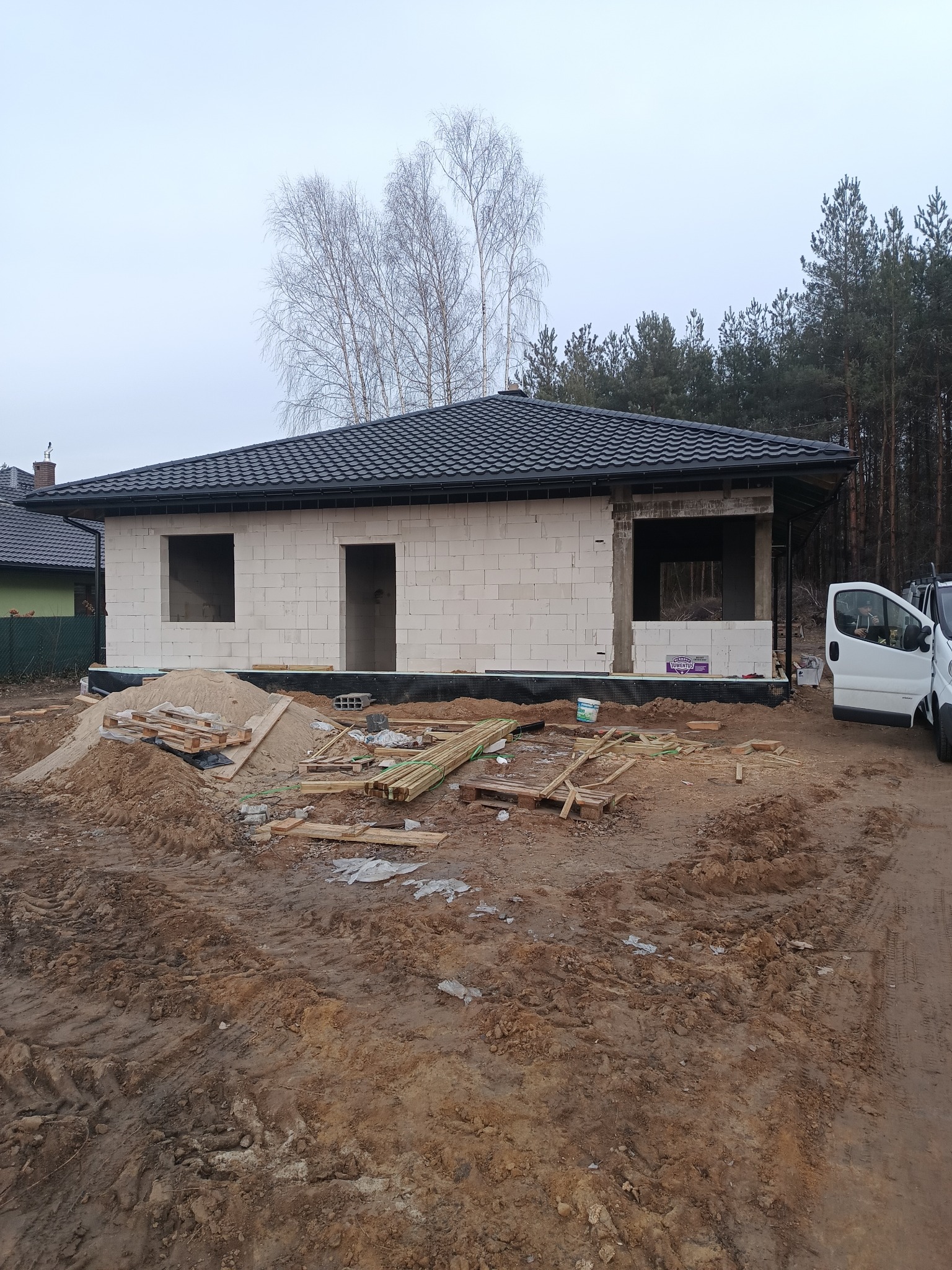 Budowa parterowego domu z białego bloczka, czarny dach z dachówki, widoczne otwory okienne i drzwiowe, teren budowy, las w tle.