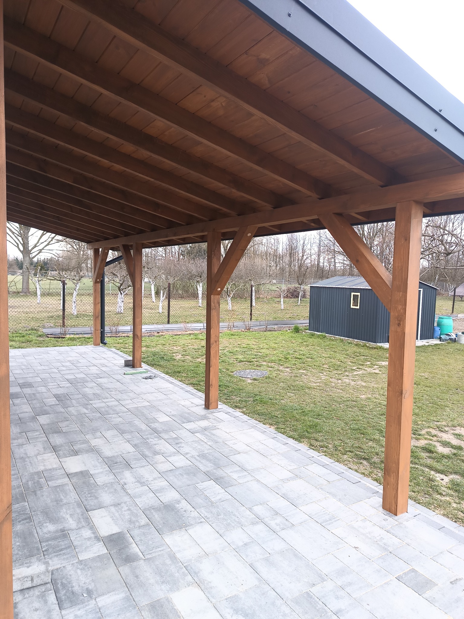 Drewniana altana z szarą, płaską podłogą z kostki brukowej. Widok na ogród z metalowym domkiem narzędziowym w tle. Konstrukcja z belek w ciepłym odcieniu brązu.