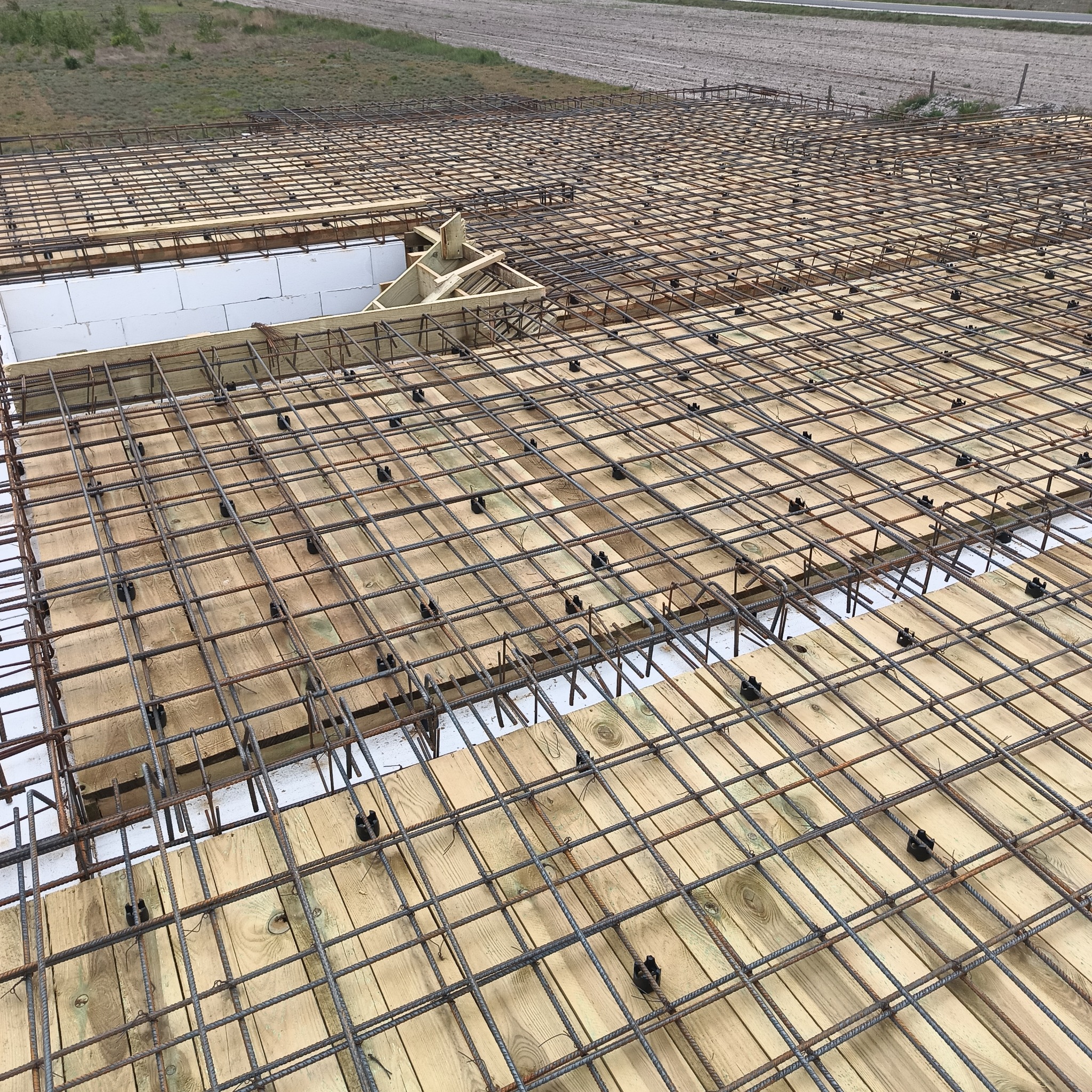 Zbrojenie stropu budynku z drewnianym szalunkiem i białymi bloczkami, widoczne z góry. Siatka zbrojeniowa ułożona na deskach, czarne elementy dystansowe.