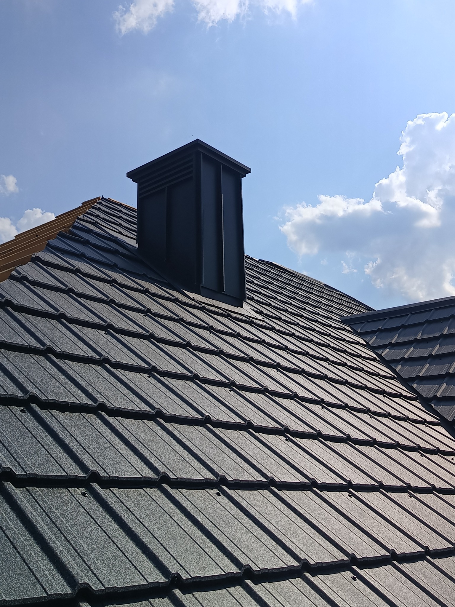 Grafitowy dach z blachodachówki modułowej i komin wentylacyjny na tle błękitnego nieba z chmurami. Widoczne mocowania blachy.