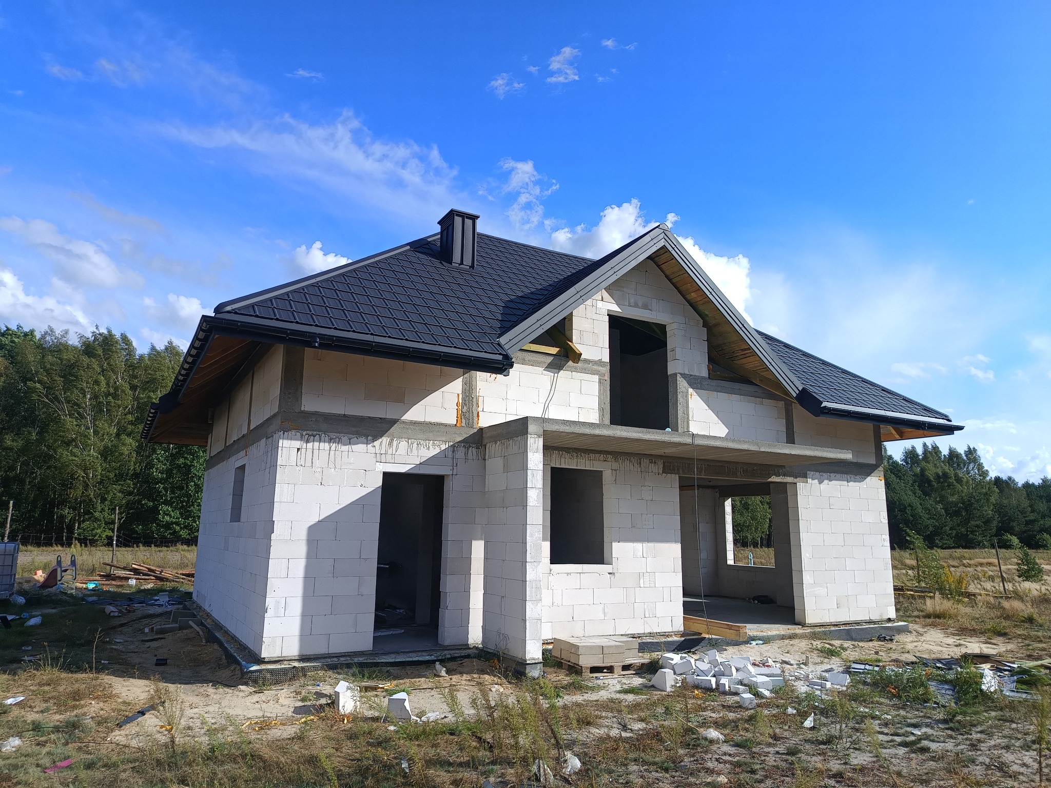 Surowy dom z białego pustaka pod czarnym dachem na tle błękitnego nieba z chmurami. Widoczne niezabudowane otwory okienne i drzwiowe. Etap budowy.