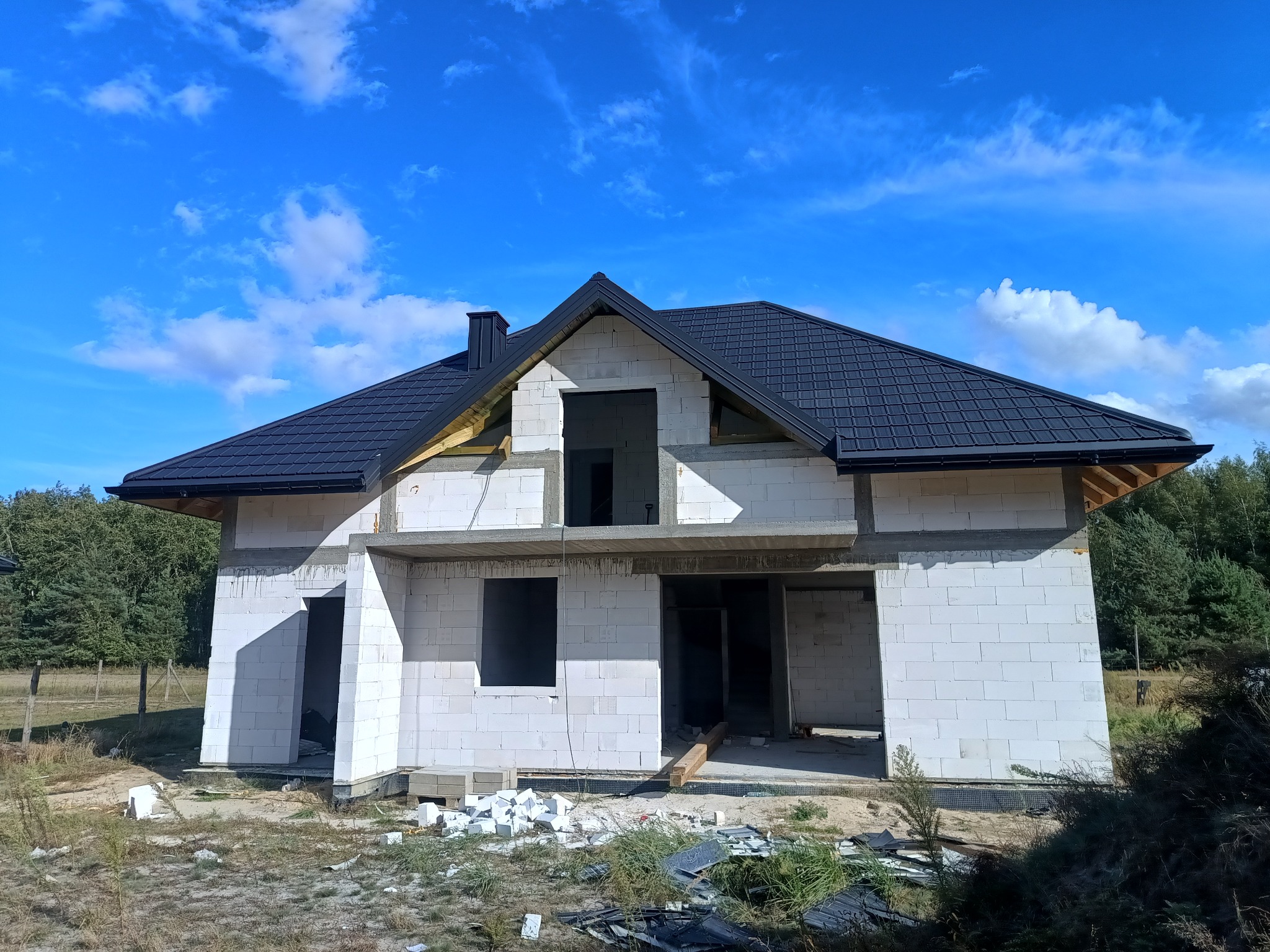 Budowa domu z białego bloczka w Godzianowie, z czarnym dachem i widocznym kominem. Stan surowy otwarty, błękitne niebo w tle. Widoczne resztki materiałów budowlanych.