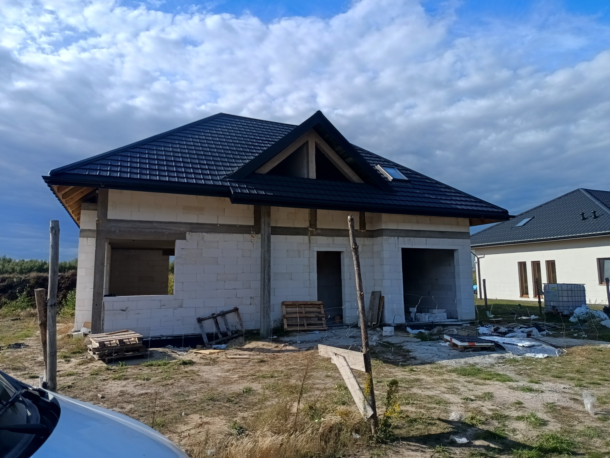 Niedokończony dom z białych bloczków i czarnym dachem, widoczne elementy konstrukcyjne, palety i gruz budowlany na zewnątrz, błękitne niebo w tle.