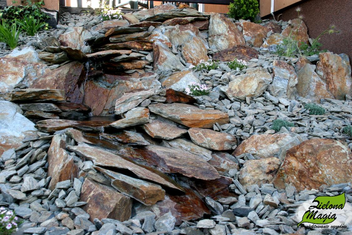 Kaskada z nieregularnych, płaskich kamieni w ogrodzie, z delikatnym przepływem wody i roślinnością w tle. Dekoracyjny element z kamienia naturalnego.