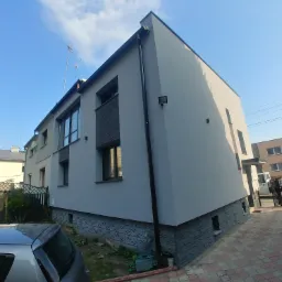 Odnowiona elewacja dwukondygnacyjnego budynku z szarą fasadą, dekoracyjnym panelowaniem i kamienną podmurówką, widoczna z perspektywy chodnika z kostki brukowej, z fragmentem zaparkowanego...