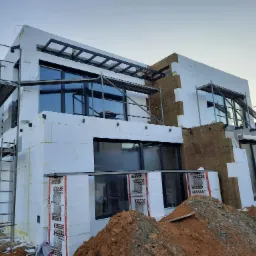 Wznoszony dwukondygnacyjny dom z elementami ocieplenia elewacji, widoczne rusztowania, styropian Besser i częściowo położona wełna mineralna, na pierwszym planie pryzma ziemi.