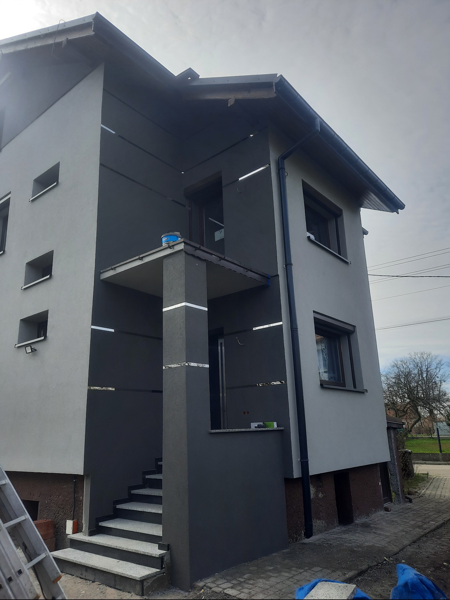 Dwukondygnacyjny dom jednorodzinny z szarą elewacją, widoczne fragmenty rusztowania, niezakończone prace wykończeniowe przy schodach wejściowych i elewacji, na balkonie stoi wiadro z farbą.