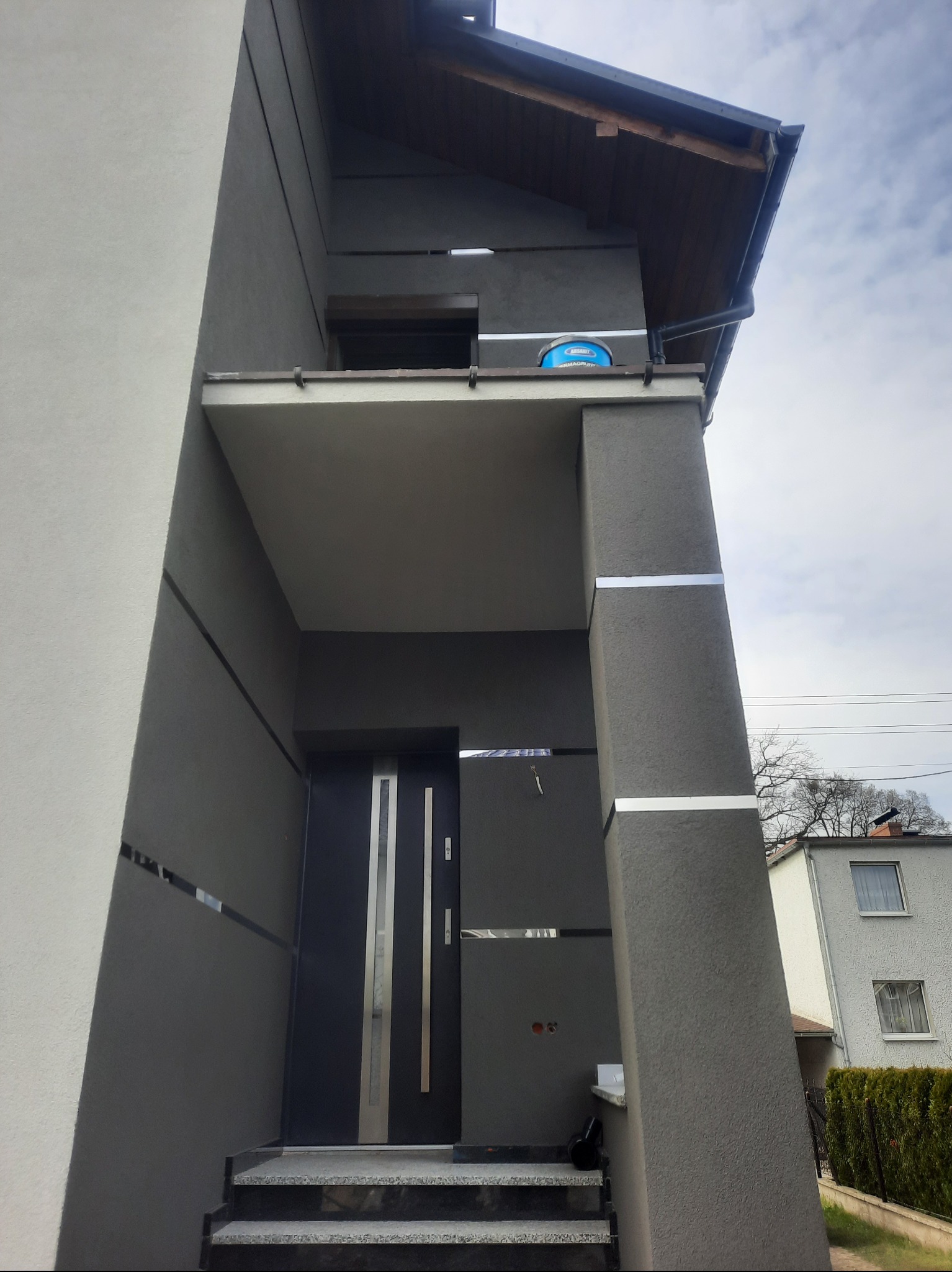 Nowoczesna elewacja domu w odcieniach szarości z elementami dekoracyjnymi w postaci poziomych, srebrnych pasków, widoczna puszka z farbą na balkonie.