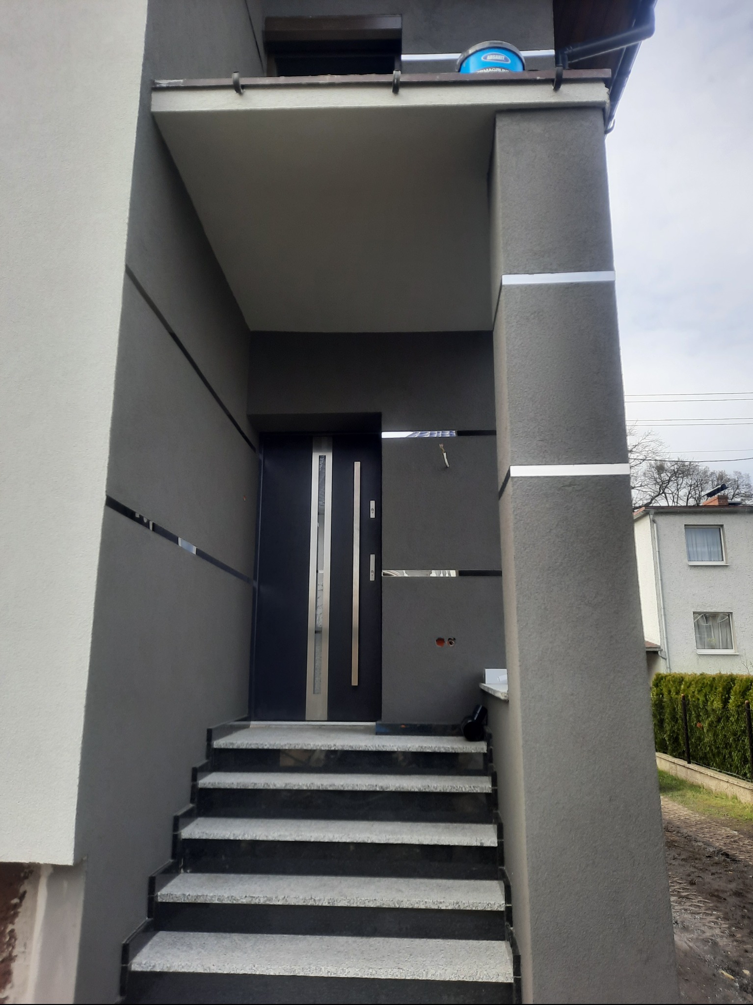 Szare wejście do domu z nowoczesnymi schodami z szarego kamienia, elewacją w odcieniach szarości z poziomymi srebrnymi akcentami i niebieskim wiadrem na dachu.