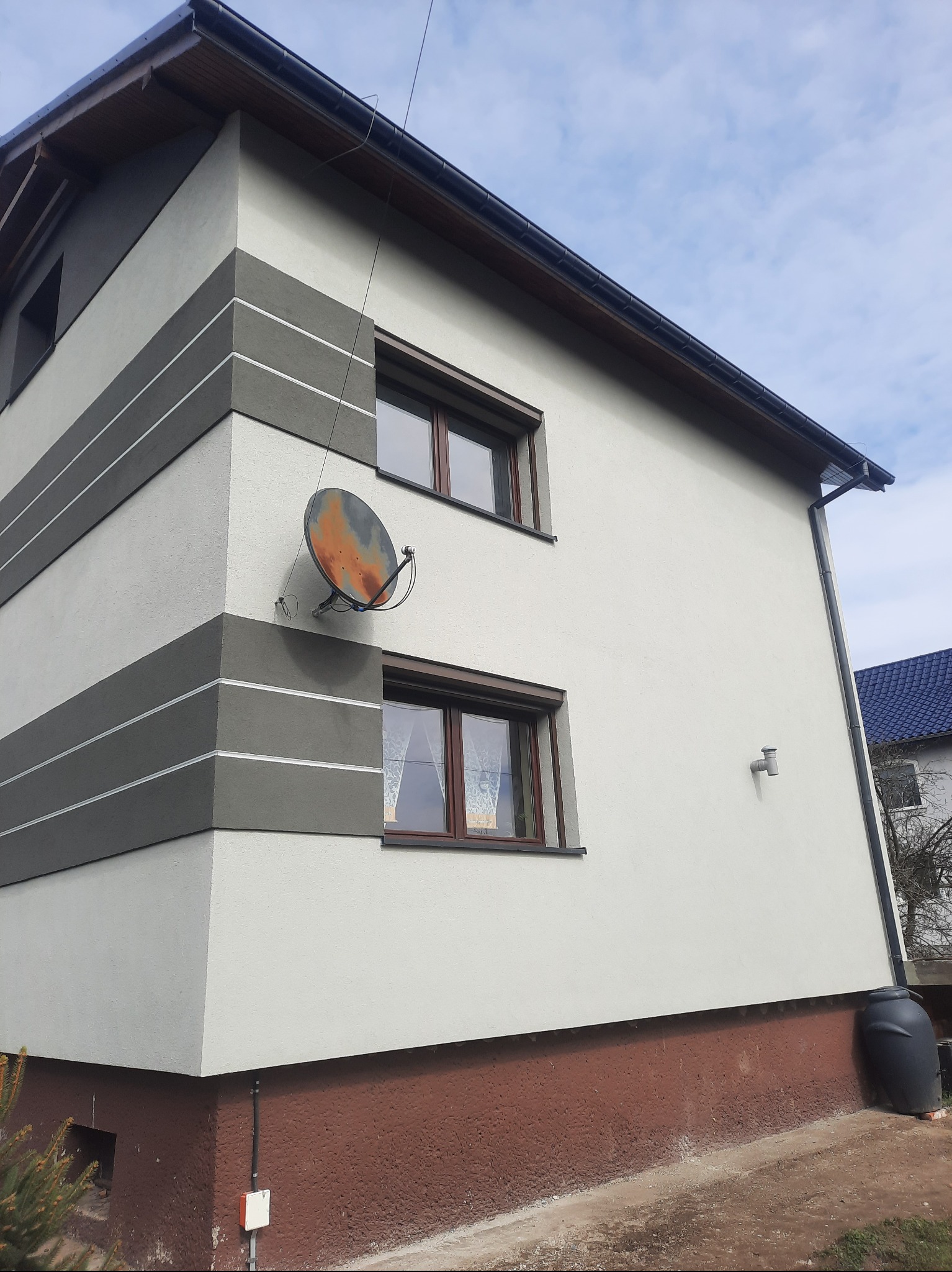 Dwukondygnacyjny dom z jasną elewacją, ciemnymi narożnikami z poziomymi paskami, brązowymi oknami i zardzewiałą anteną satelitarną na bocznej ścianie.