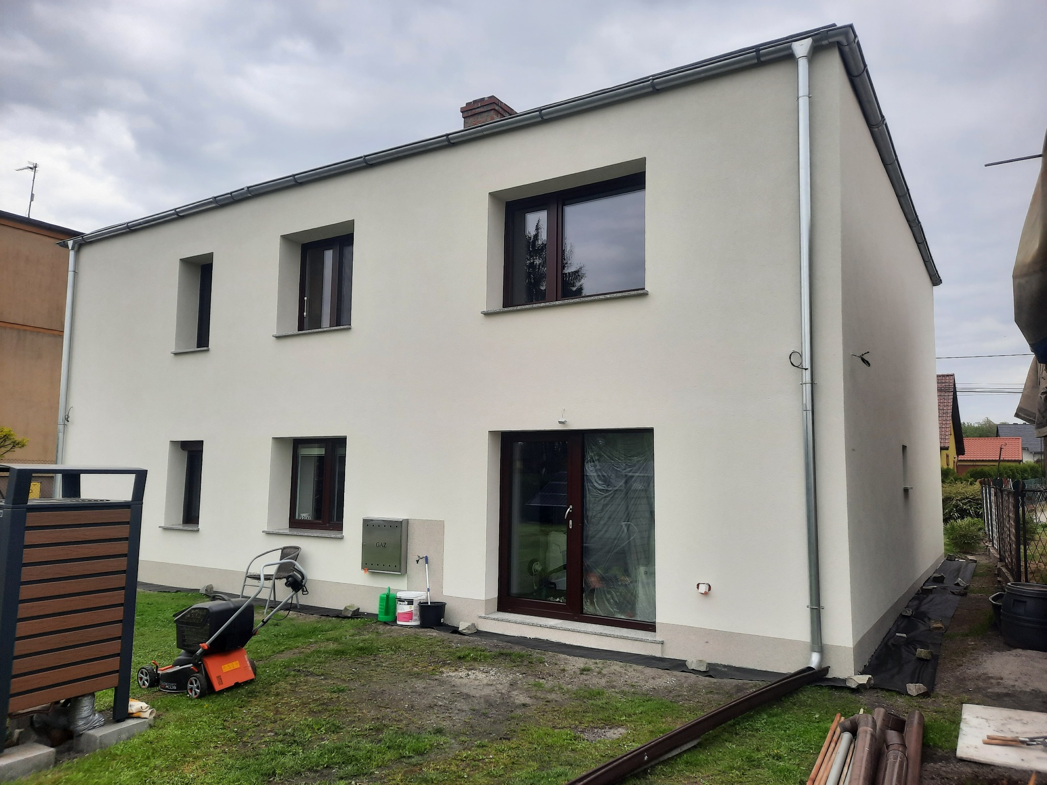 Nowoczesny, dwukondygnacyjny dom z jasną elewacją, ciemnymi oknami i metalowymi rynnami, widoczny trawnik z kosiarką i elementami przygotowawczymi do prac ziemnych.