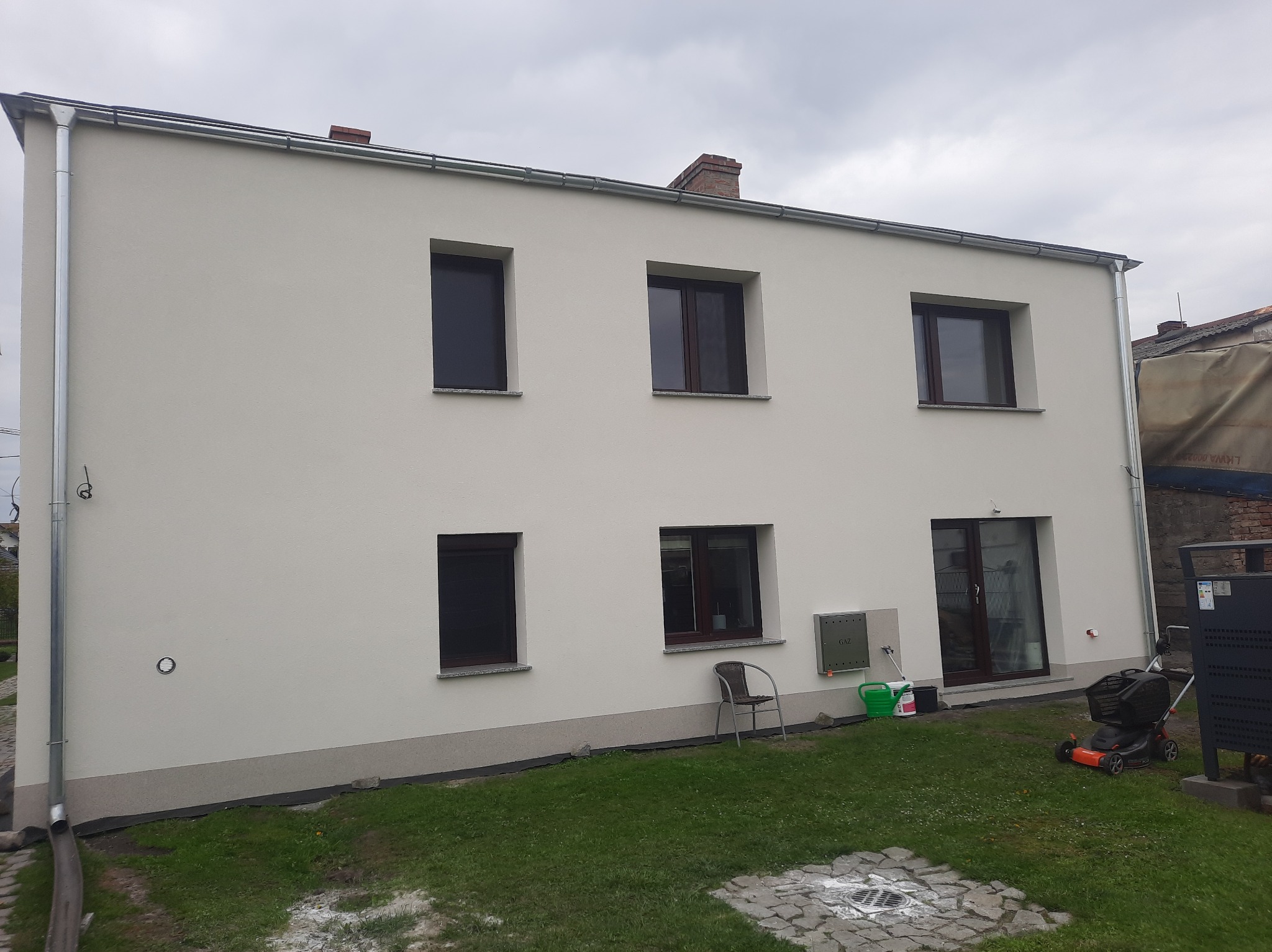 Dwukondygnacyjny dom z jasną elewacją, brązowymi oknami, rynnami i fragmentem trawnika z kosiarką, krzesłem i pojemnikami na farby.