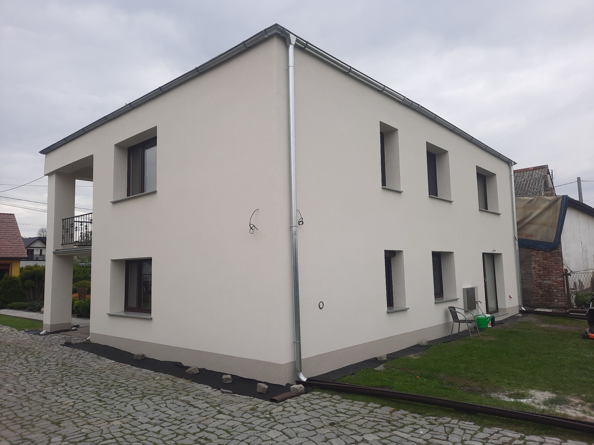 Dwukondygnacyjny dom z jasną elewacją, brązowymi oknami i balkonem, widok z poziomu kostki brukowej, z widocznymi elementami wykończeniowymi wokół budynku.