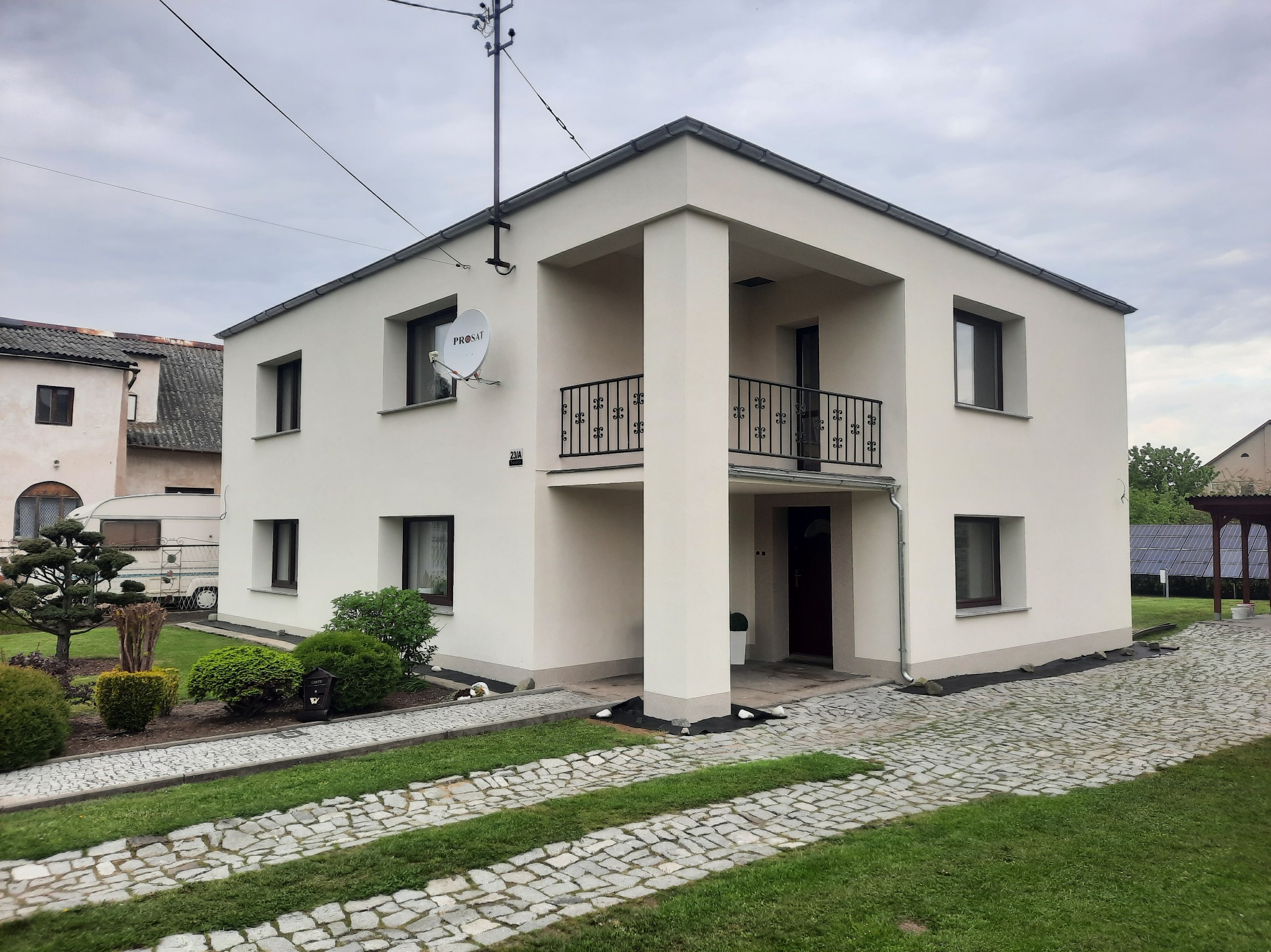 Dwukondygnacyjny dom z płaskim dachem i balkonem, elewacja w odcieniach bieli, podjazd z kostki brukowej oddzielony pasami trawy, widoczna instalacja satelitarna na ścianie.