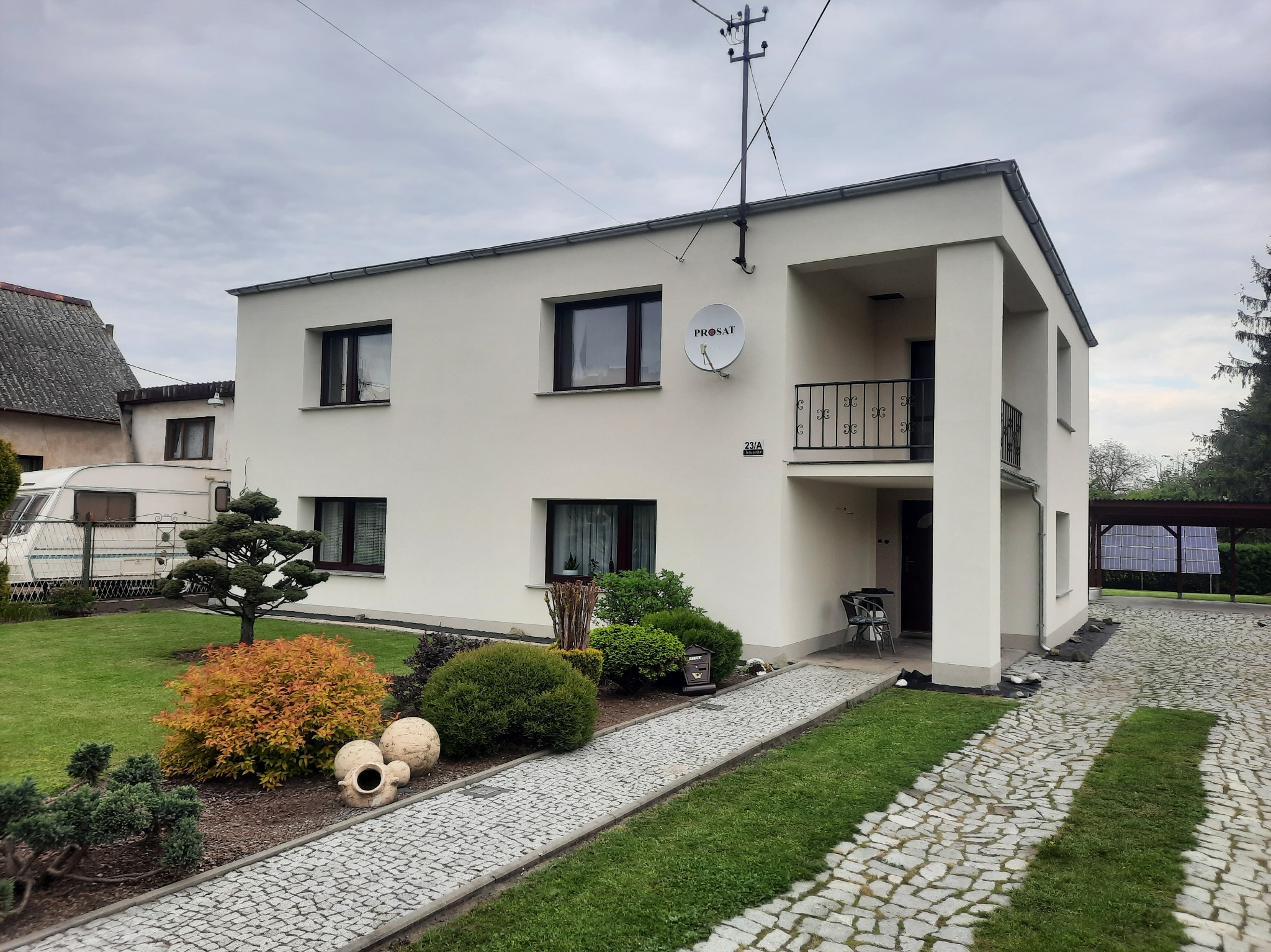 Dwukondygnacyjny dom z płaskim dachem, otynkowany na biało, z balkonem, anteną satelitarną i brukowaną ścieżką w ogrodzie.