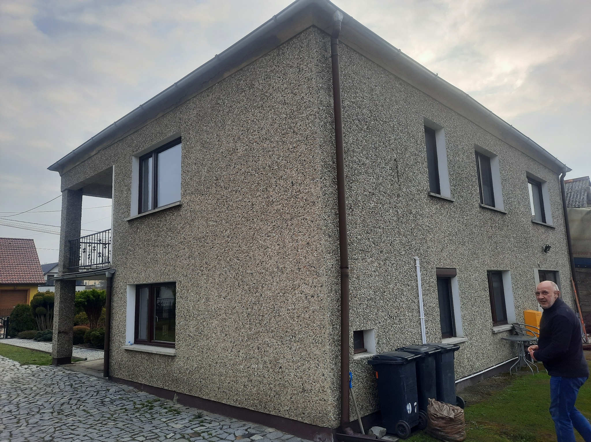 Dwupiętrowy dom z elewacją typu baranek w odcieniach szarości, widok z zewnątrz, z balkonem i trzema czarnymi pojemnikami na śmieci przy ścianie, na tle pochmurnego nieba i sąsiednich budynków.