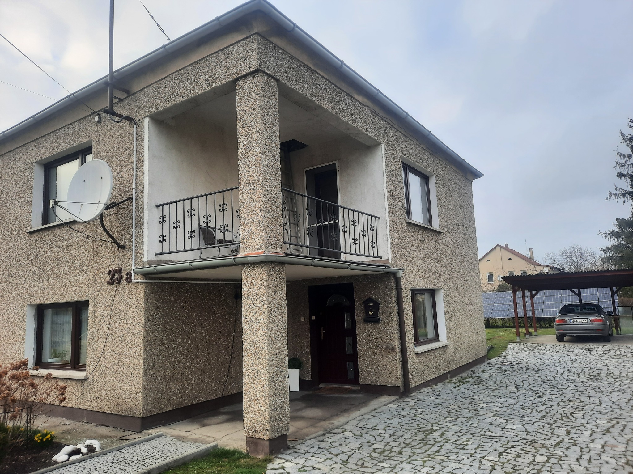 Dwupiętrowy dom z elewacją o strukturze baranka, balkonem z kutą balustradą i podjazdem z kostki brukowej, w tle wiata garażowa z panelami słonecznymi.