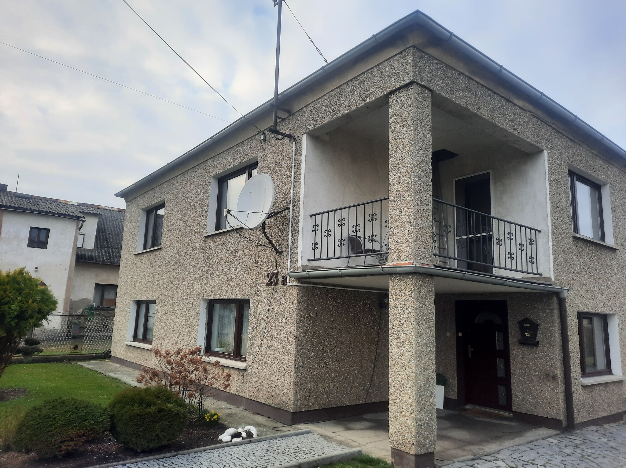 Dwupiętrowy dom z elewacją typu baranek, balkonem z kutą balustradą i anteną satelitarną przy oknie.