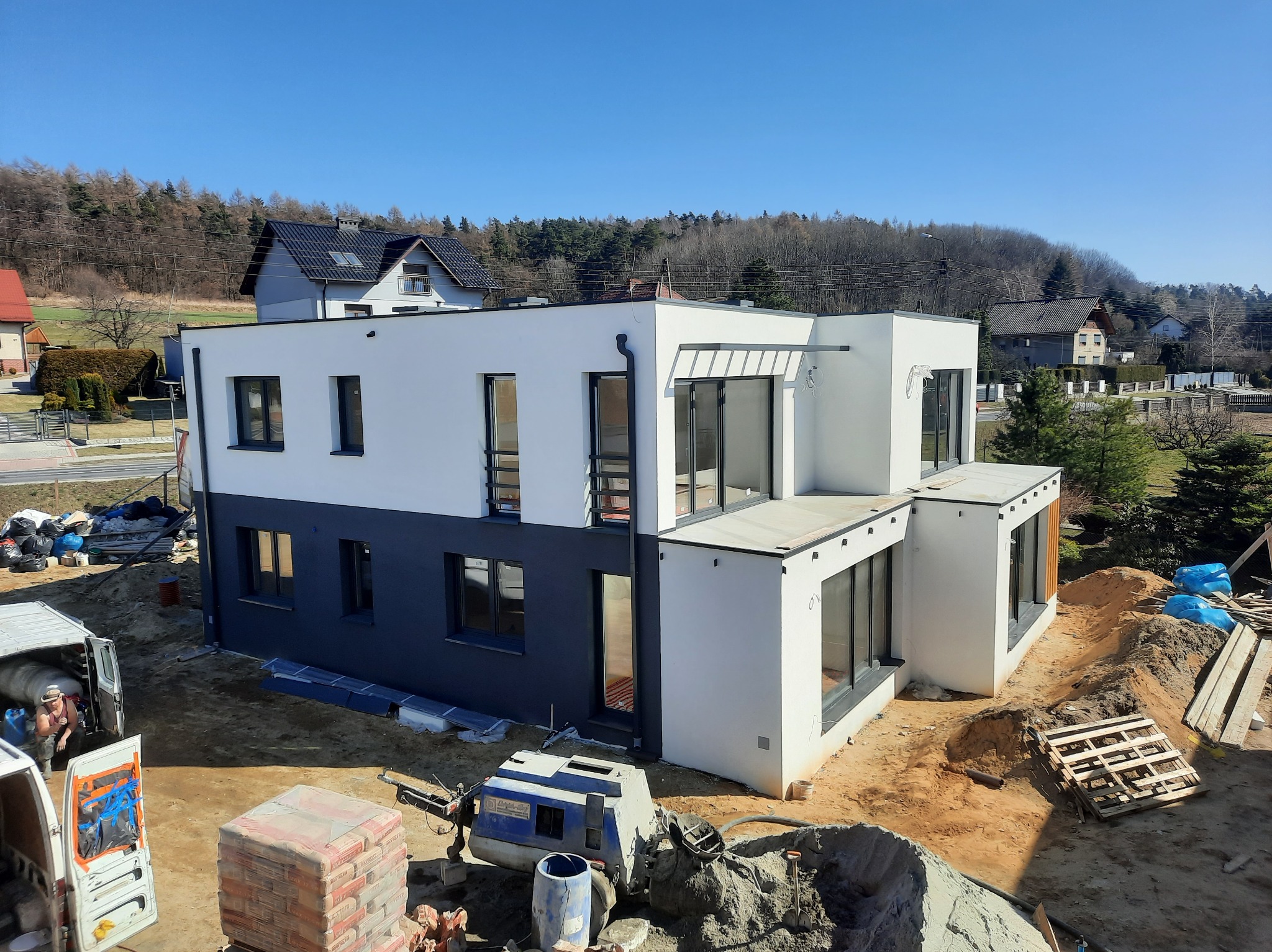 Dwukondygnacyjny dom w trakcie budowy, widoczne worki z cementem, betoniarka i pracownik w samochodzie dostawczym, w tle inne budynki i drzewa.