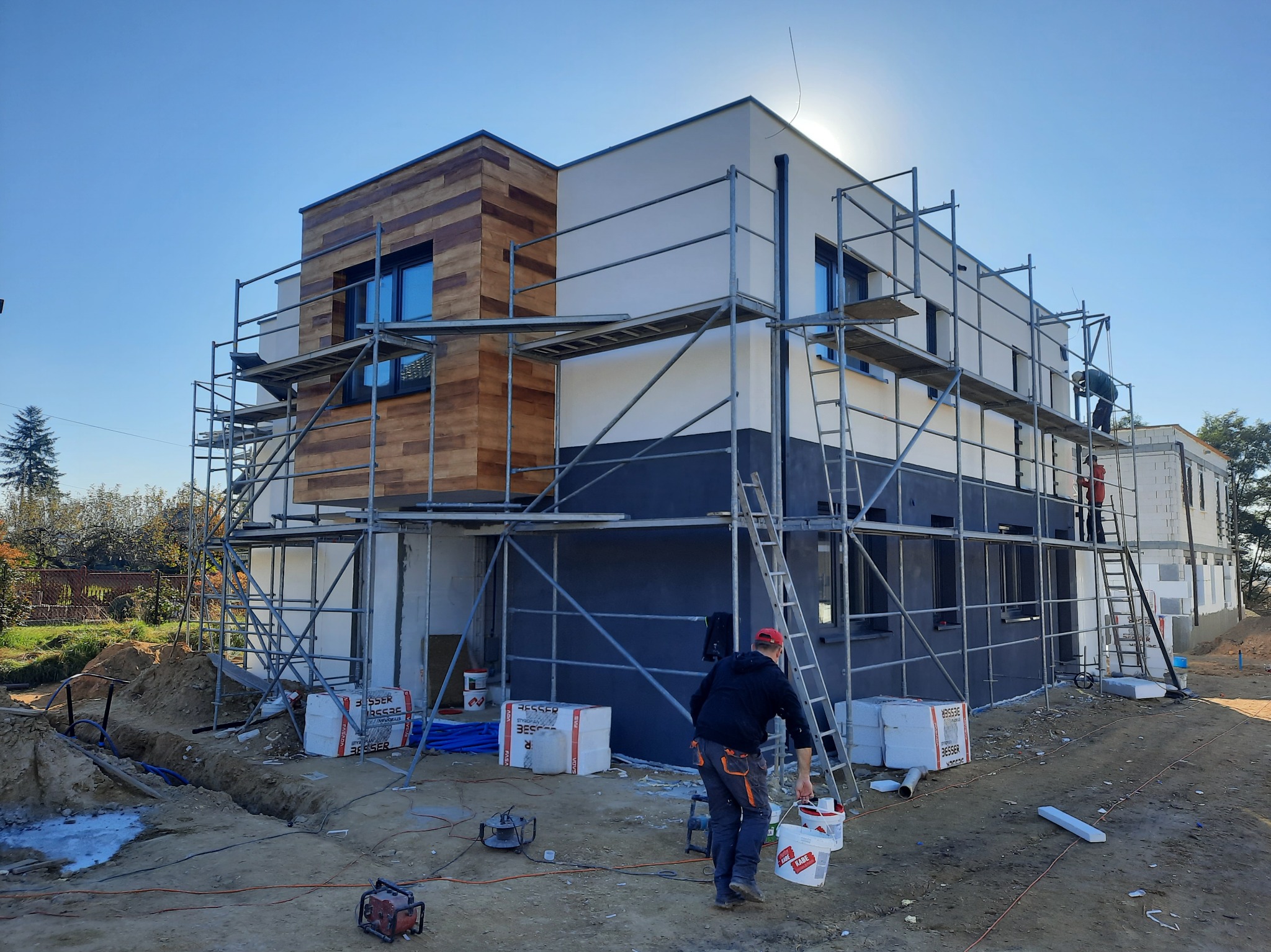 Nowoczesny dom dwukondygnacyjny w trakcie prac elewacyjnych, częściowo wykończony drewnianą okładziną, otoczony rusztowaniami i materiałami budowlanymi, z pracownikami na górze i jednym niosącym...