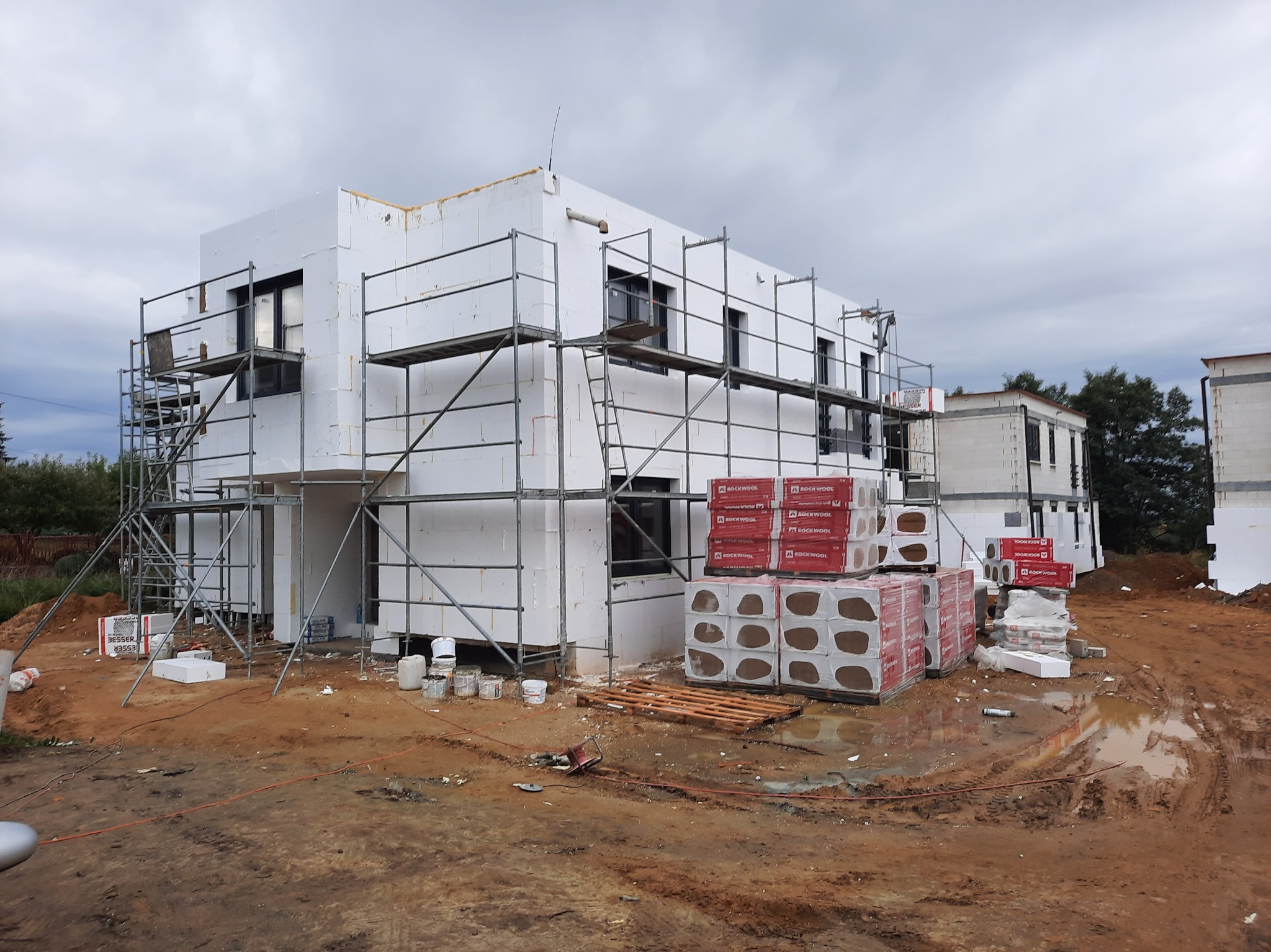 Dwukondygnacyjny dom w trakcie ocieplania styropianem, widoczne rusztowania, palety z materiałem izolacyjnym Rockwool, teren budowy błotnisty po deszczu, pochmurne niebo.