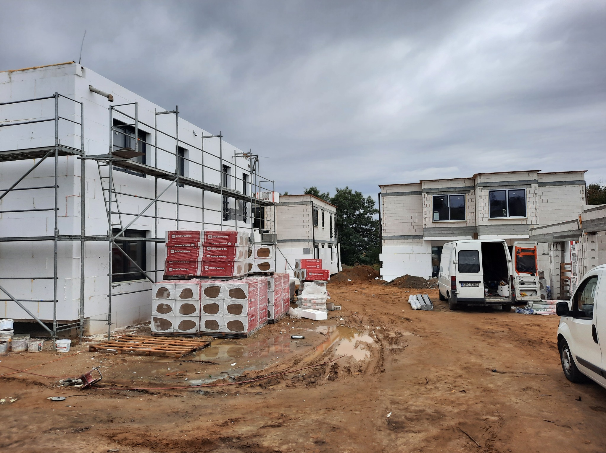 Budowa szeregowych domów z białego styropianu, widoczne rusztowania, palety z materiałami izolacyjnymi Rockwool, teren budowy z błotem i śladami opon