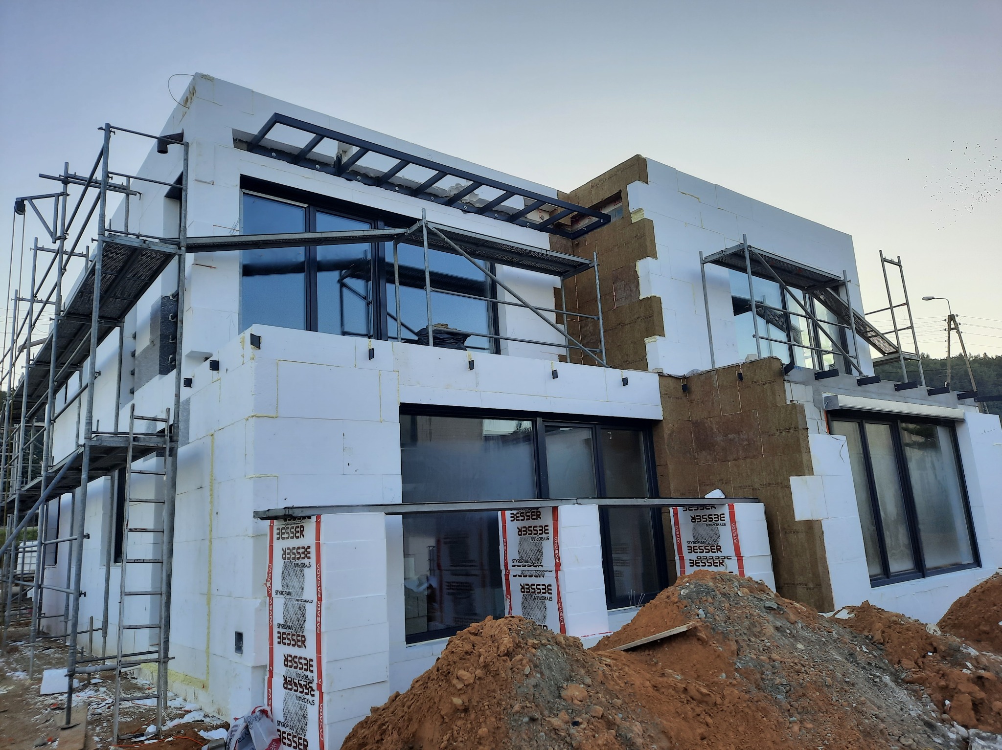 Wznoszony dwukondygnacyjny dom z elementami ocieplenia elewacji, widoczne rusztowania, styropian Besser i częściowo położona wełna mineralna, na pierwszym planie pryzma ziemi.
