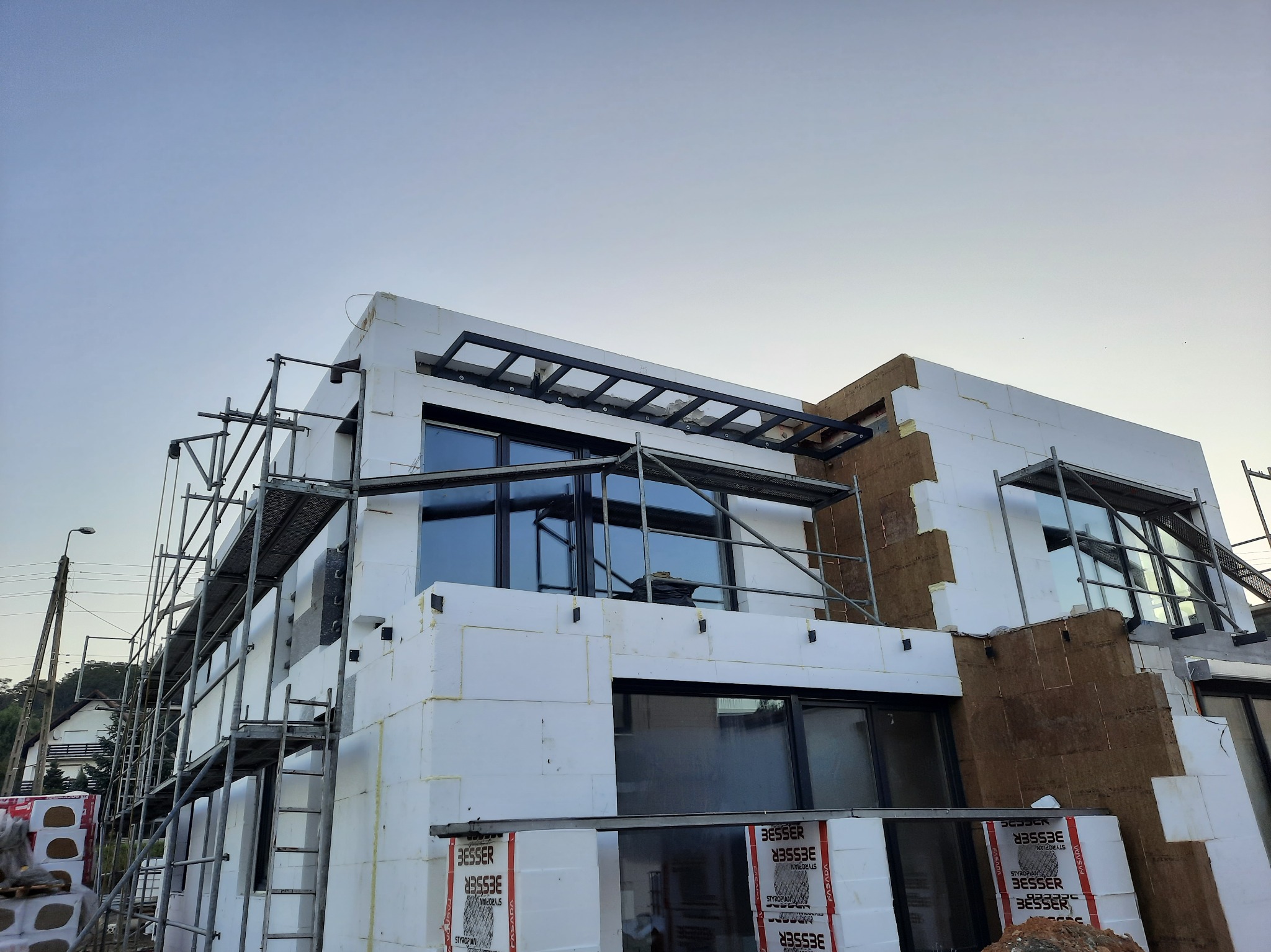 Nowoczesny dom w trakcie ocieplania elewacji, widoczne rusztowania, styropian i częściowo ocieplona ściana z brązową wełną mineralną, w tle słup energetyczny i zabudowa jednorodzinna.