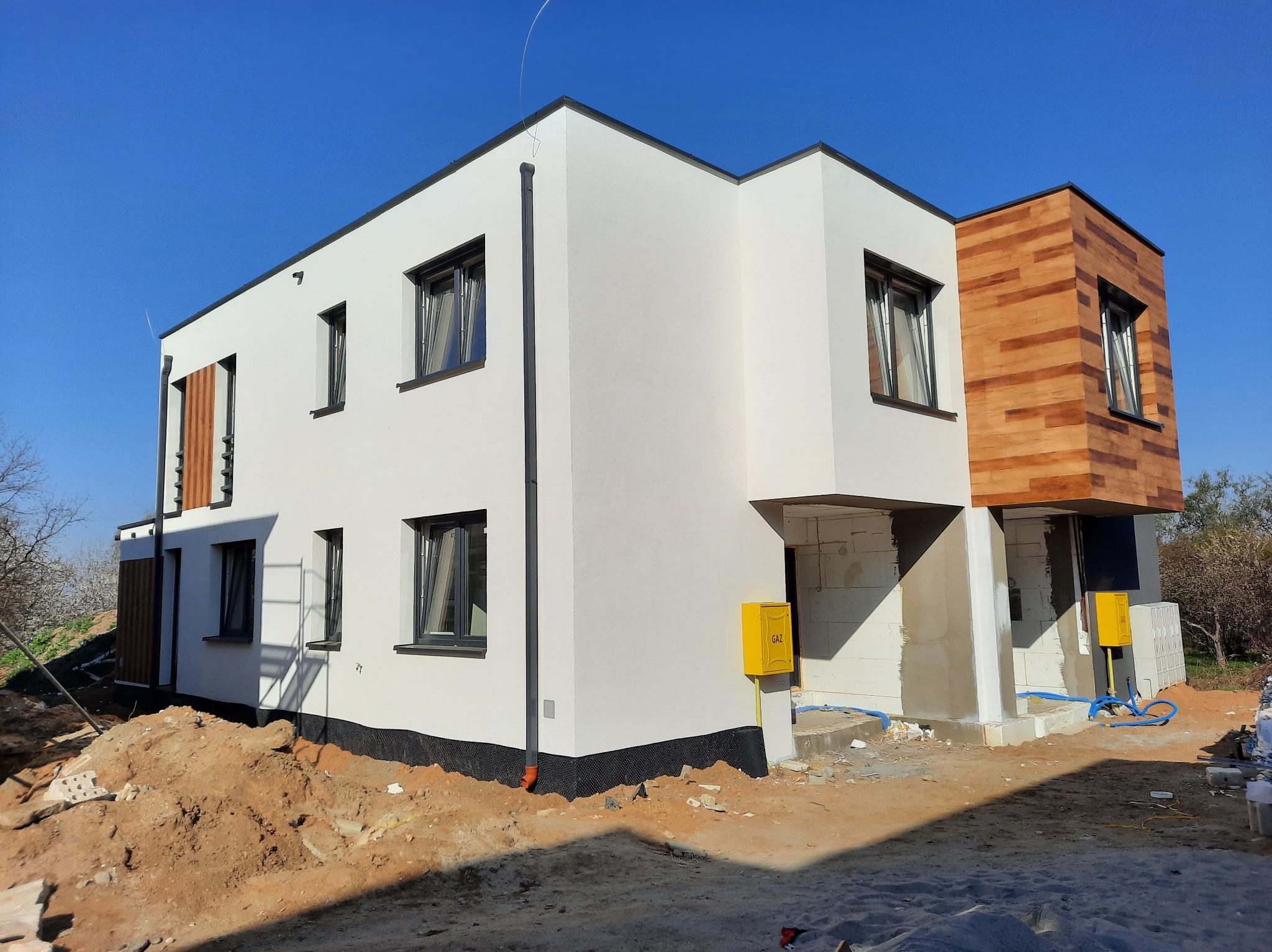 Nowoczesny, dwukondygnacyjny dom w trakcie budowy z elementami elewacji wykończonymi drewnem, widoczny żółty licznik gazu na elewacji oraz przygotowany teren wokół budynku.