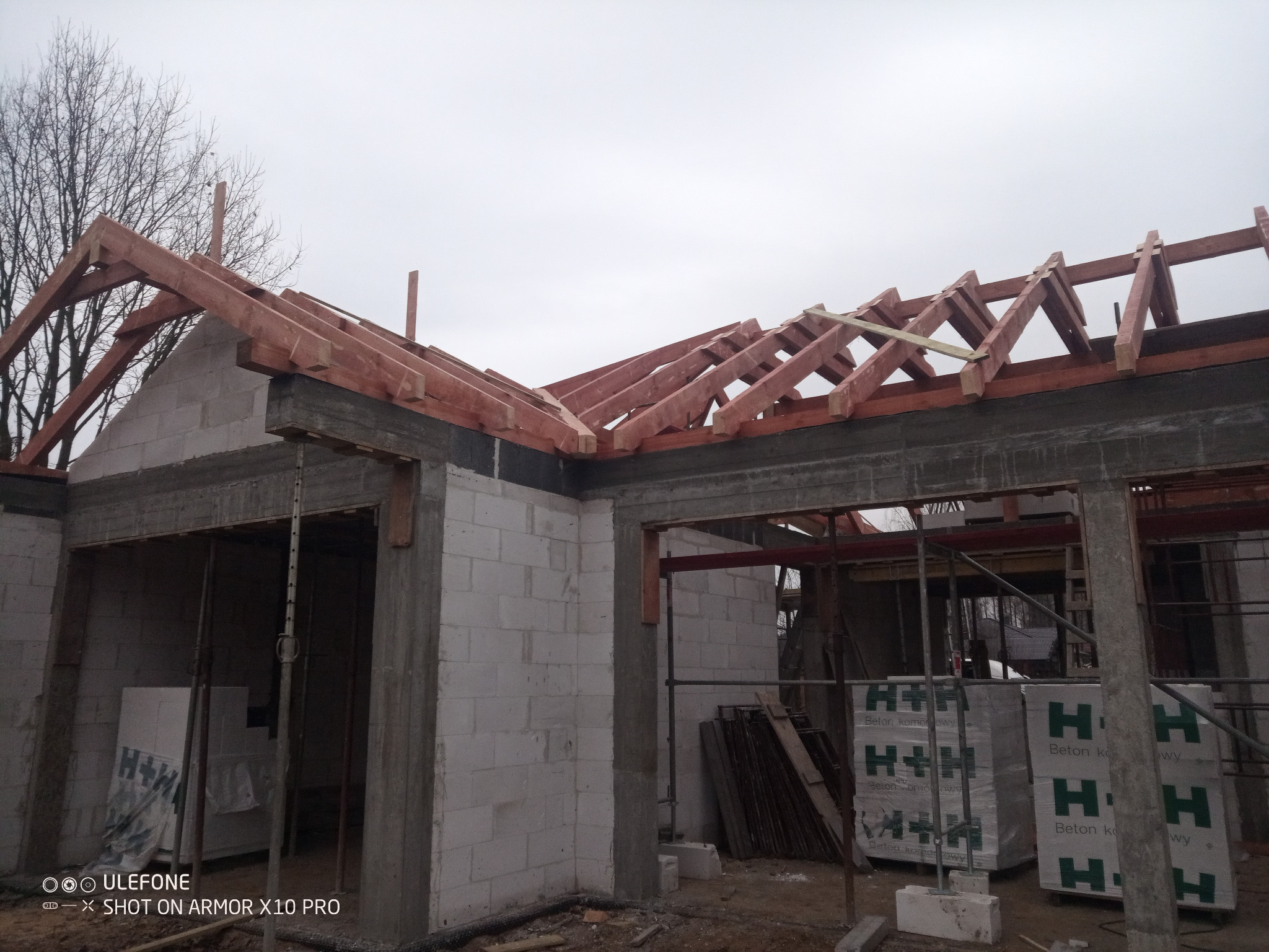 Surowa konstrukcja domu z widoczną drewnianą więźbą dachową w trakcie budowy. Elementy konstrukcyjne z betonu komórkowego. Szary, pochmurny dzień.