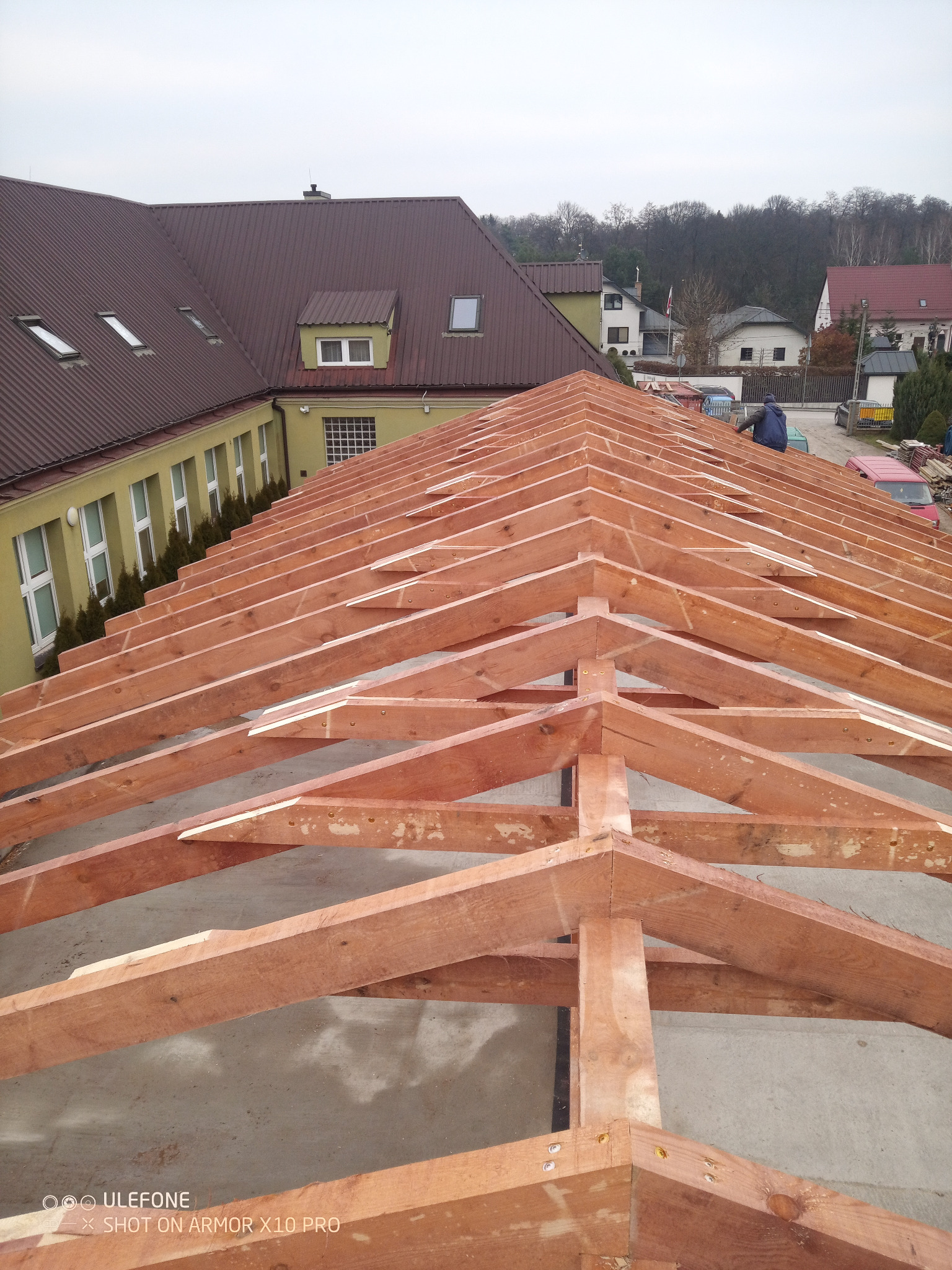 Konstrukcja drewniana więźby dachowej w trakcie montażu, widok z góry, z fragmentem budynku w tle. Widoczny pracownik na dachu.