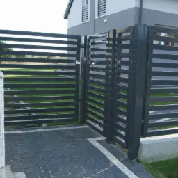 Nowoczesna, grafitowa brama wjazdowa z poziomymi panelami, otwierana na posesję wyłożoną kostką brukową. Widoczny fragment domu w tle i skrzynka elektryczna.