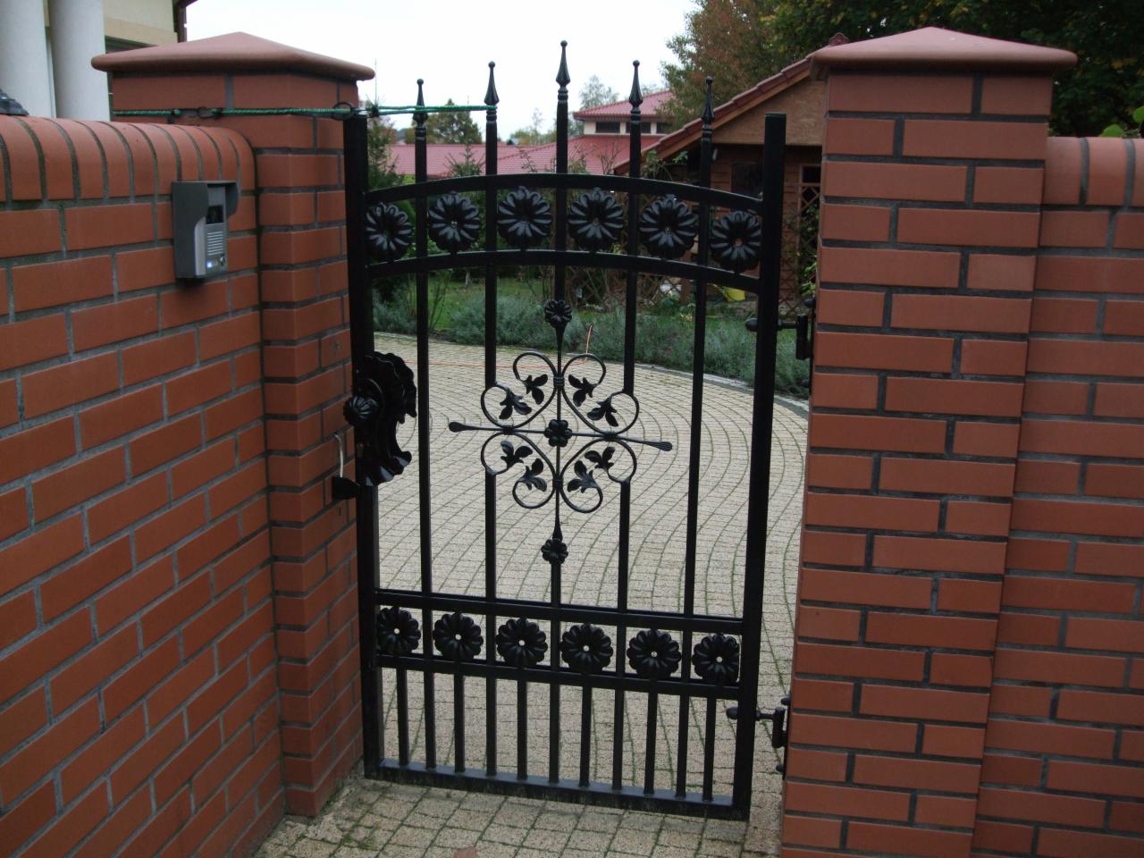 Otwarta, czarna, kuta furtka z motywem kwiatowym, umieszczona między dwoma ceglanymi filarami. Widoczny dom w tle i kostka brukowa.