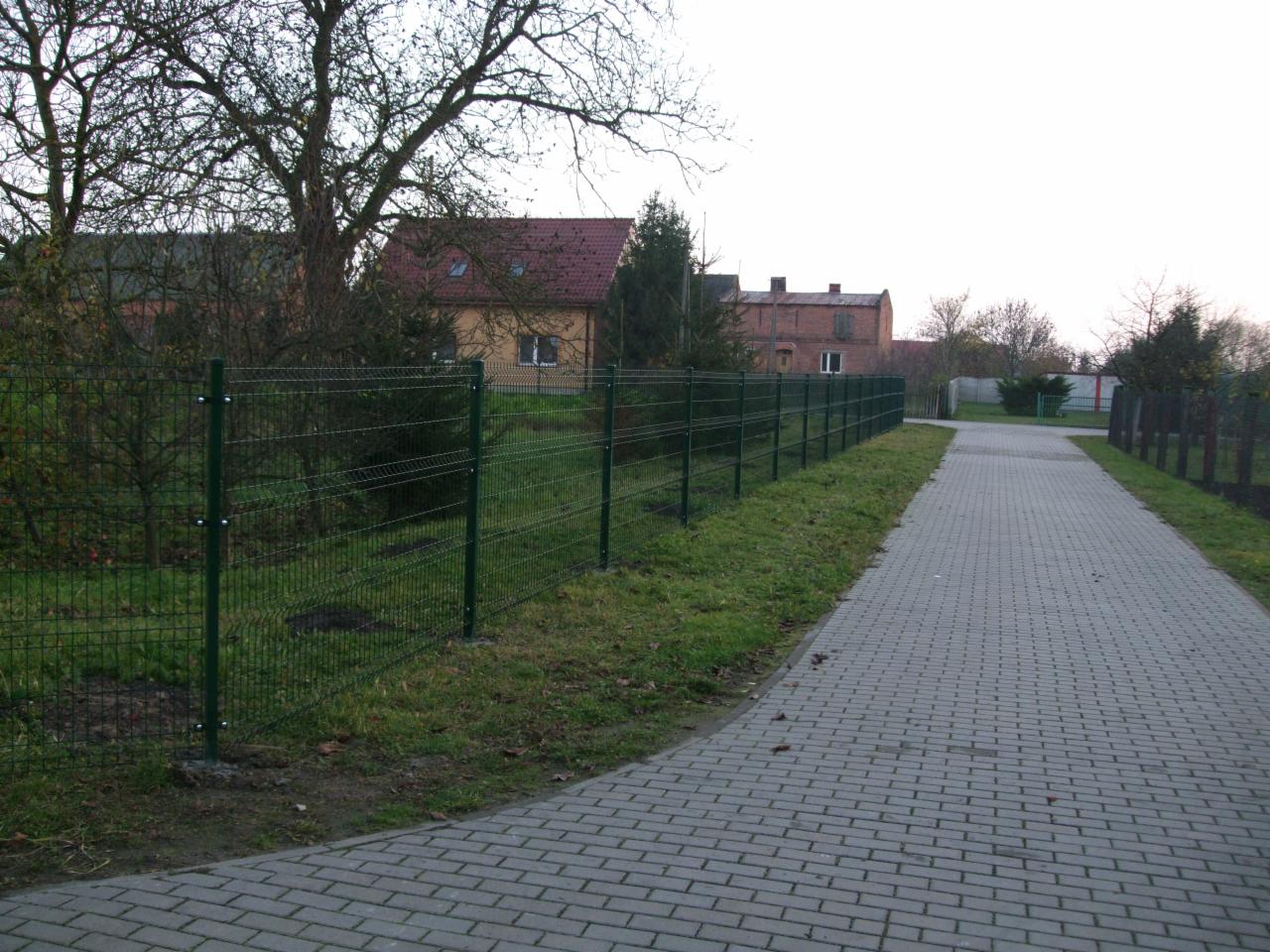 Długi, zielony panel ogrodzeniowy z widocznymi śrubami, oddzielający posesję od ścieżki z kostki brukowej. W tle budynki i drzewa.