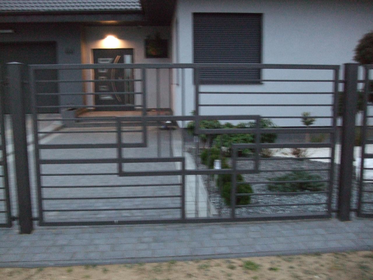 Nowoczesna, metalowa brama wjazdowa z geometrycznym wzorem, na tle domu z szarymi elewacjami i kostką brukową. Widoczne nasadzenia wewnątrz posesji.