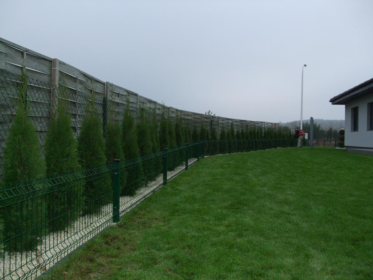 Długie, zielone ogrodzenie panelowe z siatką na podmurówce z białego kamienia, oddzielające trawnik od drewnianego płotu. Osoba w oddali przy budynku.