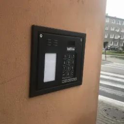 Czarny panel domofonu Laskomex z klawiaturą numeryczną, zamontowany na pomarańczowej ścianie budynku mieszkalnego, widok z boku.