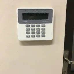 Panel sterowania alarmu Satel Perfecta zamontowany na ścianie obok drzwi z metalową klamką, wyświetlający aktualną datę i godzinę.